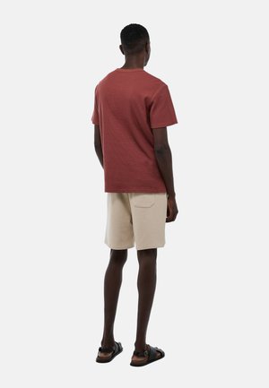 T-shirt rouge à manches courtes à côtes, shorts beiges et sandales noires. Le T-shirt a un col rond ; les shorts comportent une poche arrière et une coupe décontractée.