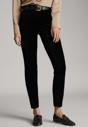 Woman wearing black fitted pants with a black belt, beige long-sleeve shirt, and dark brown loafers standing against a plain background.