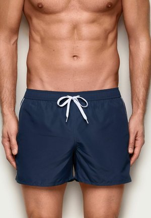Torsó masculino con bañador azul marino con cordón blanco, mostrando un abdomen tonificado en forma y brazos relajados a los lados.