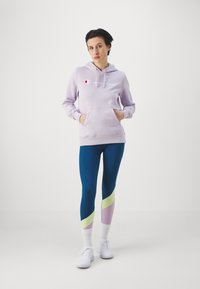 Felpa con cappuccio lavanda con tasca frontale e testo bianco "Champion", abbinata a leggings blu con accenti verdi e rosa. Sneakers bianche.