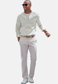 Hellgelbes und weiß gestreiftes Hemd mit Knöpfen, cremefarbene Chinos, weiße Turnschuhe und dunkle Sonnenbrille, die lässig mit den Händen in den Taschen dasteht.