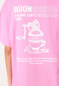 Rosa t-skjorte med hvit grafisk design av en kaffetrakter og kopp, sammen med italiensk tekst om å nyte kaffe. Mykt stoff, løs passform.