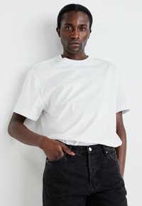 Tricou alb din bumbac, cu o croială relaxată, guler rotund, mâneci scurte, asortat cu blugi negri din denim, cu sistem de închidere cu buton și buzunare.