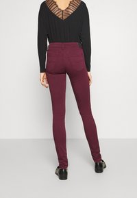 Jeans skinny burdeos de cintura alta con bolsillos traseros, combinados con una blusa negra que cuenta con un diseño de espalda transparente y rayas.