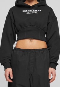 Svart kortärmad hoodie med huva och långa ärmar, prydd med vit broderad text "KARL KANI EST. 1989" på framsidan och ribbad fåll.
