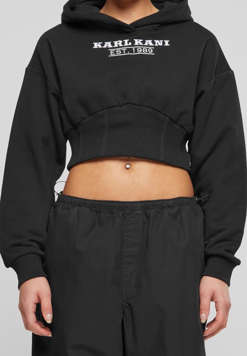 Svart kortärmad hoodie med huva och långa ärmar, prydd med vit broderad text "KARL KANI EST. 1989" på framsidan och ribbad fåll.
