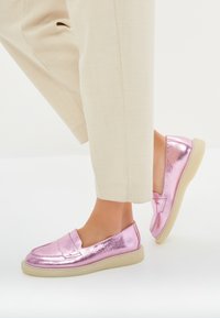 Vaaleanpunaiset metallinhohtoiset loaferit, joissa on sileä pinta, pyöristetty kärki ja vaaleanvärinen pohja, yhdistettynä beigeihin housuihin.
