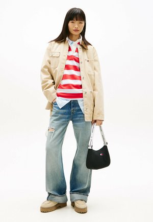 Mujer joven con chaqueta beige, camiseta de rayas rojas y blancas, vaqueros anchos rotos, zapatos beige, sosteniendo un bolso negro con correa blanca.