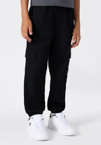 Pantaloni cargo neri con tasche laterali, polsini elasticizzati e vestibilità rilassata, abbinati a sneakers bianche con dettagli verdi.