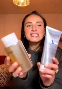 Femme aux cheveux foncés tient et montre deux produits pour les soins des cheveux, l'un dans une bouteille transparente et l'autre dans un tube argenté, dans une pièce chaleureusement éclairée.