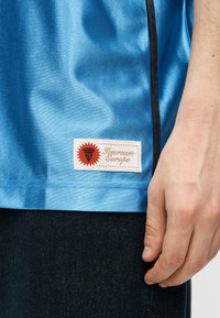 Χέρι που ακουμπά στο πλάι, φορώντας ανοιχτό μπλε πουκάμισο με μαύρη επένδυση και έμβλημα "Icecream Europe" πάνω από σκούρο παντελόνι.