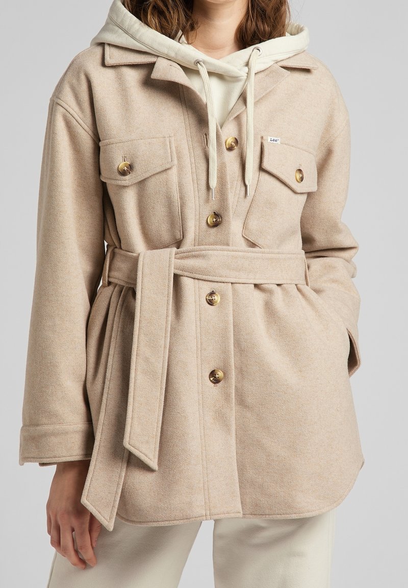 Mujer vestida con una chaqueta de lana beige con cinturón y grandes botones, sobre una sudadera crema y pantalones crema, con la mano en el bolsillo, frente a un fondo gris.