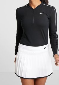 Black long-sleeve athletic top with zipper and white side stripes, paired with a white pleated skirt featuring a black hem, Nike logo accent.