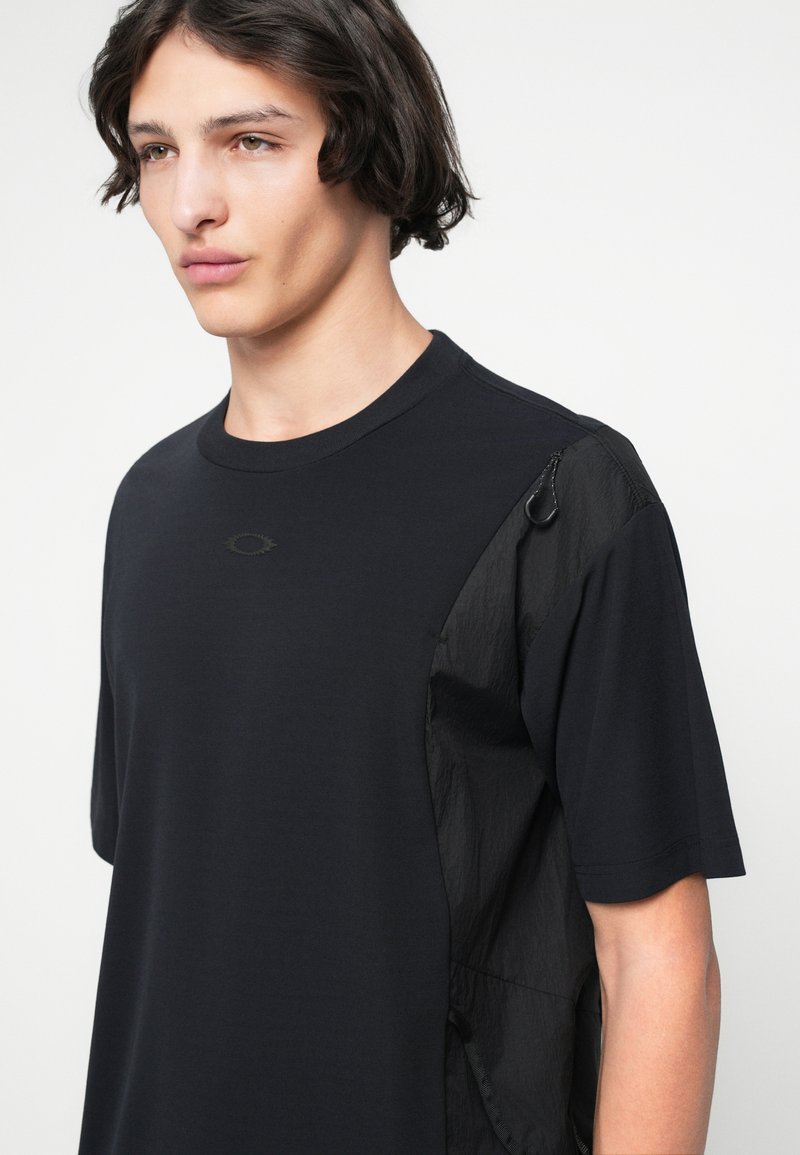 Schwarzes T-Shirt mit einem strukturierten Einsatz an der linken Schulter und an der Seite, mit einem dezenten Logo auf der Brust und einem runden Halsausschnitt.