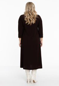 Longue robe marron à manches trois-quarts, texture lisse et coupe décontractée, associée à des bottes blanches montantes jusqu'aux genoux.