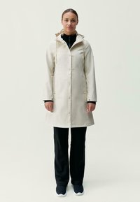 Krémovo sfarbený pršiplášť s gombíkovým zapínaním, nastaviteľnou kapucňou a bočnými vreckami, skombinovaný s čiernymi nohavicami a tmavými teniskami. Hladká textúra.