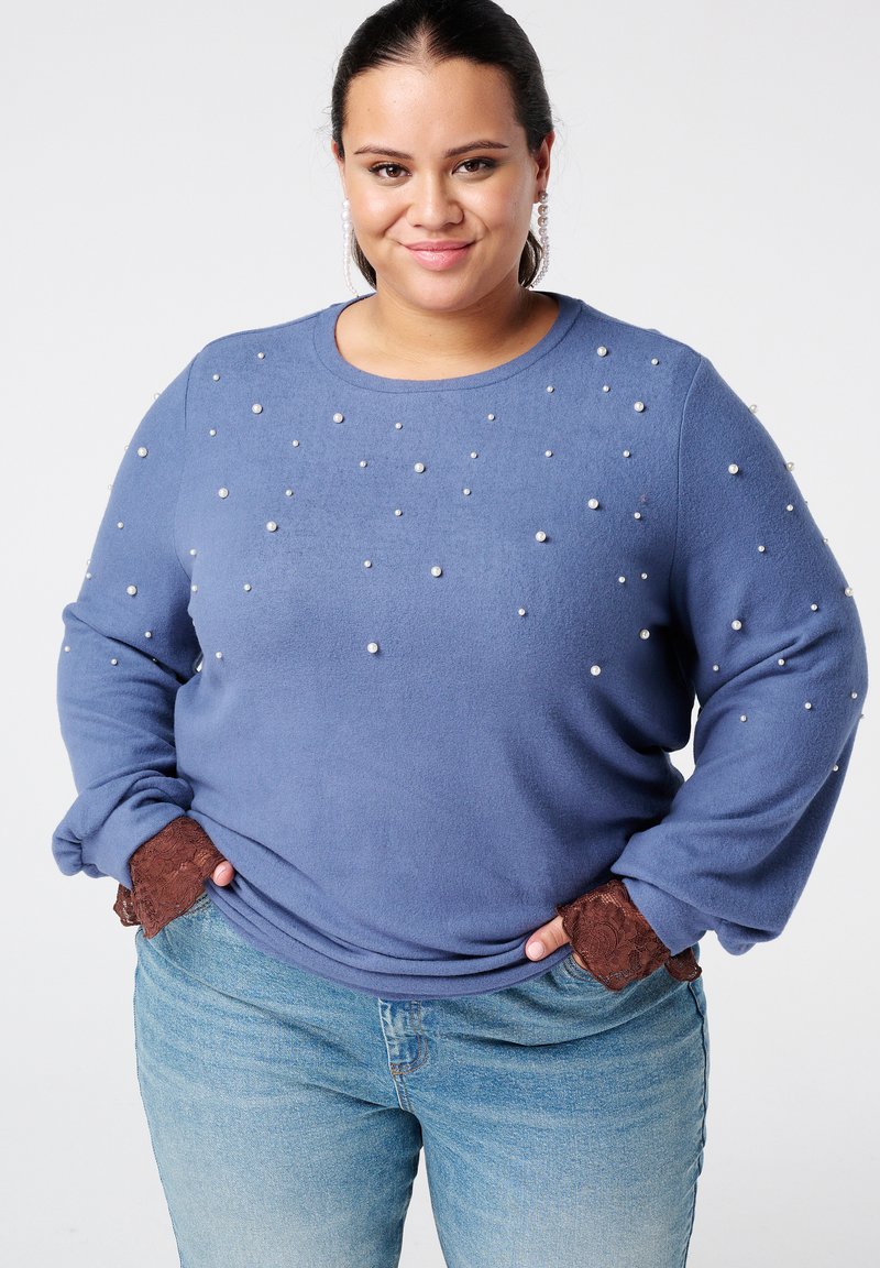 Vrouw met donker haar, gekleed in een blauwe trui versierd met kleine witte parels, kant manchetten, parel oorbellen en lichtblauwe jeans, lachend.