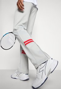 Hellgraue Sporthosen mit roten und weißen gestreiften Akzenten an den Bündchen, inklusive einem Reißverschlussdetail. Weiße Sneakers runden das Outfit ab. Ein Tennisschläger wird gehalten.
