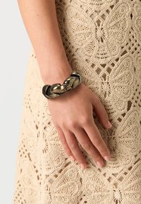 Sorte og bronse armbånd med sammenflettede lenker og en jevn metallisk finish, båret på en hånd mot en teksturert beige heklet kjole.