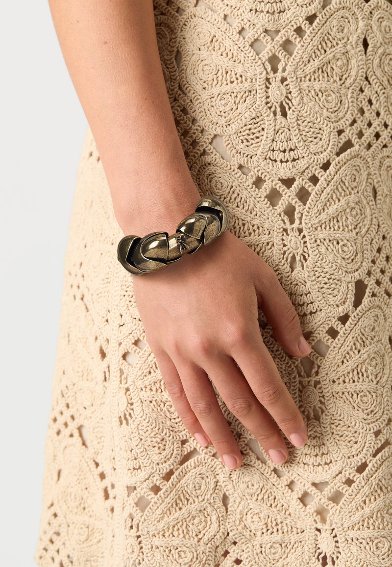 Sorte og bronse armbånd med sammenflettede lenker og en jevn metallisk finish, båret på en hånd mot en teksturert beige heklet kjole.