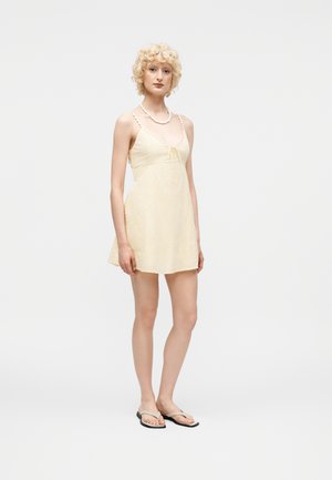 Νεαρό άτομο με σγουρά ξανθά μαλλιά, που φοράει ένα ανοιχτό κίτρινο αμάνικο φόρεμα, μαύρα σαγιονάρες και ένα λευκό κολιέ, στέκεται πάνω σε λευκό φόντο.