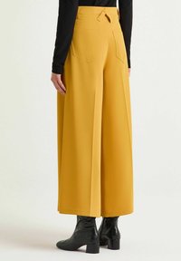 Pantaloni gialli a gamba larga con vita alta, vestibilità dritta, due tasche posteriori e una texture liscia, abbinati a stivaletti neri lucidi.