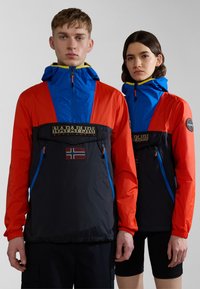 Chaquetas de pulóver coloridas en azul, naranja y negro, con capucha, cremallera y bolsillos frontales. Presenta un logo y un parche de bandera. Textura suave.