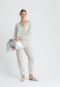 Helle Minzbluse mit V-Ausschnitt und Bindeband, kombiniert mit beigen, taillierten Hosen und metallischen, silbernen Slippern. Graue Tasche in der Hand.
