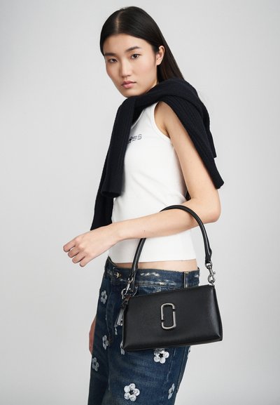 Borsa a tracolla in pelle nera con forma strutturata, caratterizzata da un dettagli in metallo argentato ben visibile. Il modello indossa una maglietta bianca e jeans blu ricamati con fiori.