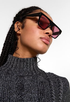Giovane donna con capelli intrecciati che indossa grandi occhiali da sole con montatura nera e lenti rosa e un maglione dolcevita grigio scuro strutturato.