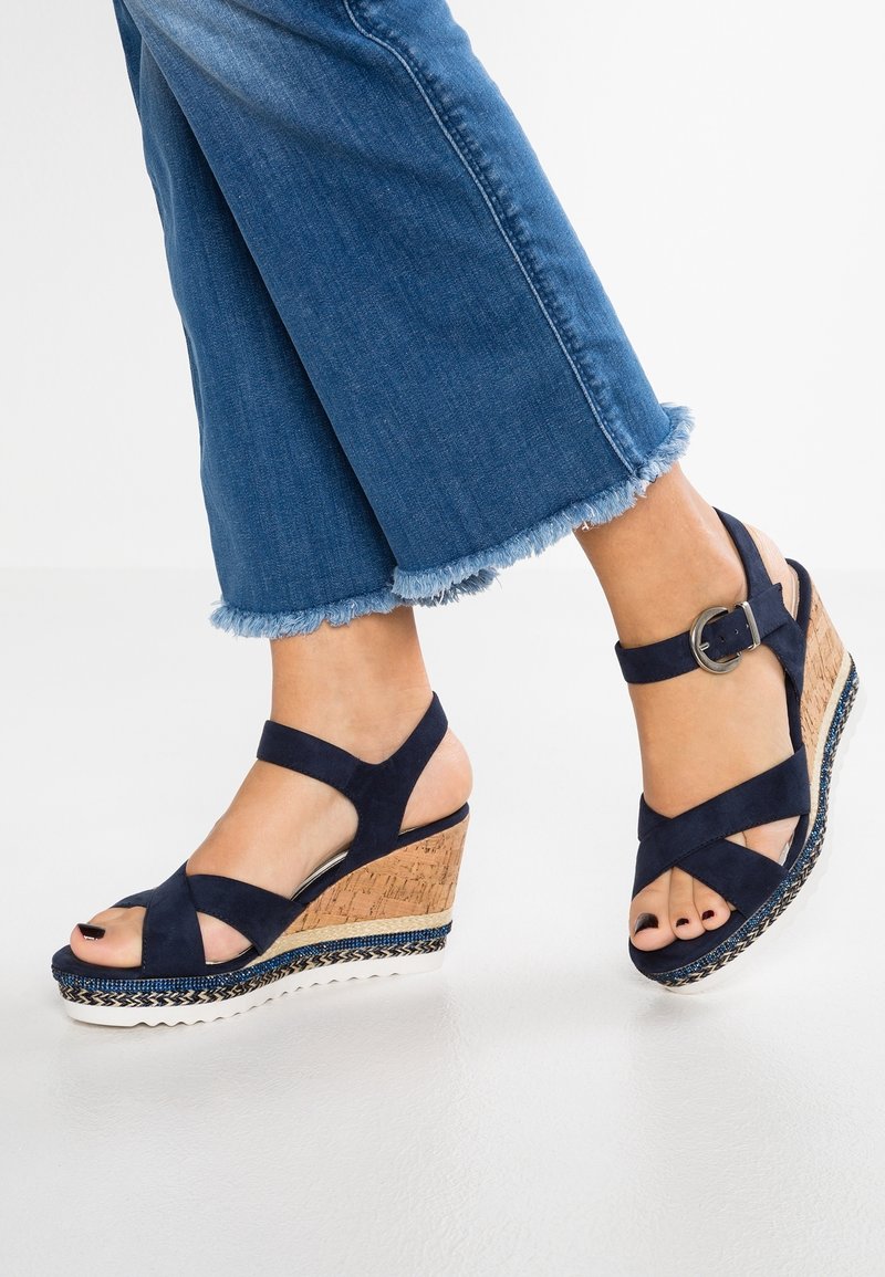 Jambes en jean bleu court avec des ourlets effilochés, portant des sandales compensées bleu marine avec des talons en liège et des brides de cheville avec des boucles argentées.