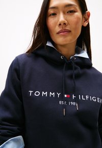 Granatowa bluza z kapturem ze sznurkiem, na której znajduje się logo "TOMMY HILFIGER" w kolorach białym, czerwonym i niebieskim. Pod spodem widoczna jasnoniebieska koszula z kołnierzykiem.