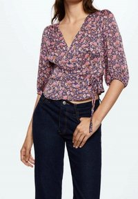 Blusa avvolgente floreale in viola con fiori rosa e bianchi, caratterizzata da una profonda scollatura a V, maniche a sbuffo e dettaglio con fiocco laterale. Abbinata a jeans scuri.