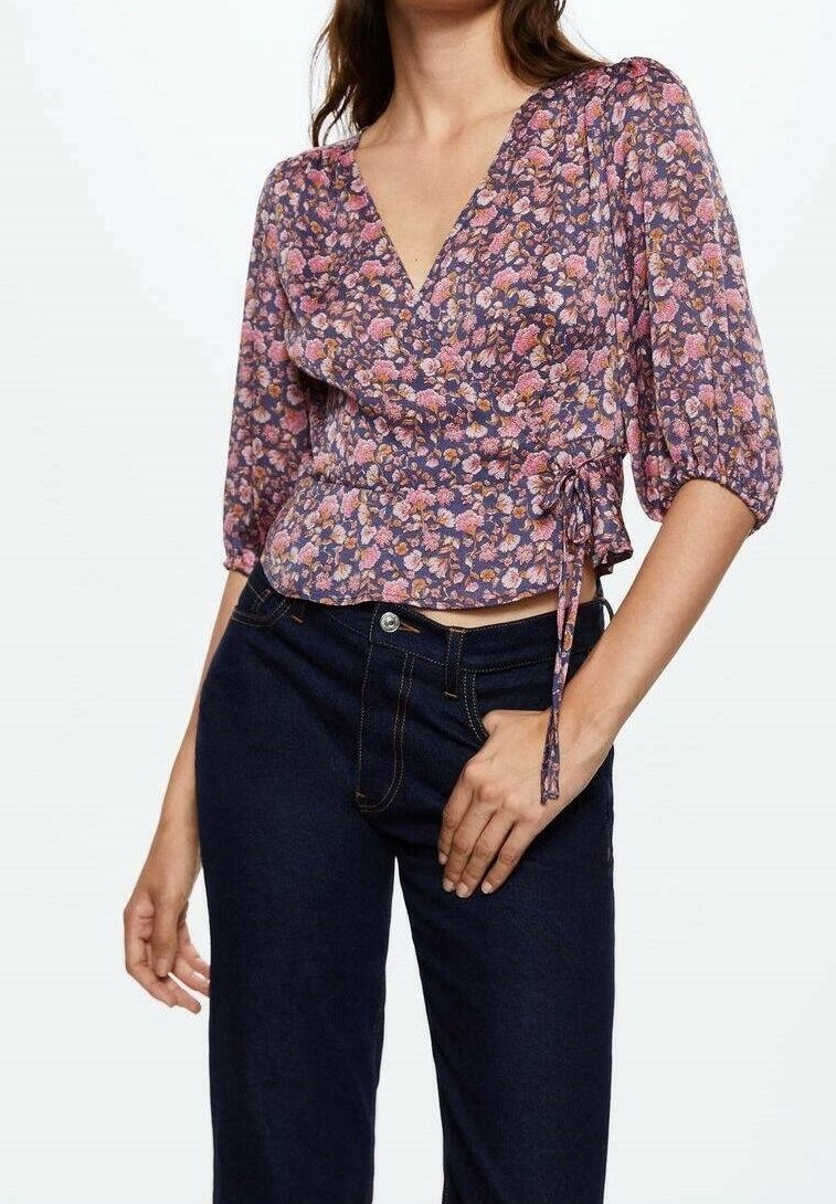 Blusa avvolgente floreale in viola con fiori rosa e bianchi, caratterizzata da una profonda scollatura a V, maniche a sbuffo e dettaglio con fiocco laterale. Abbinata a jeans scuri.