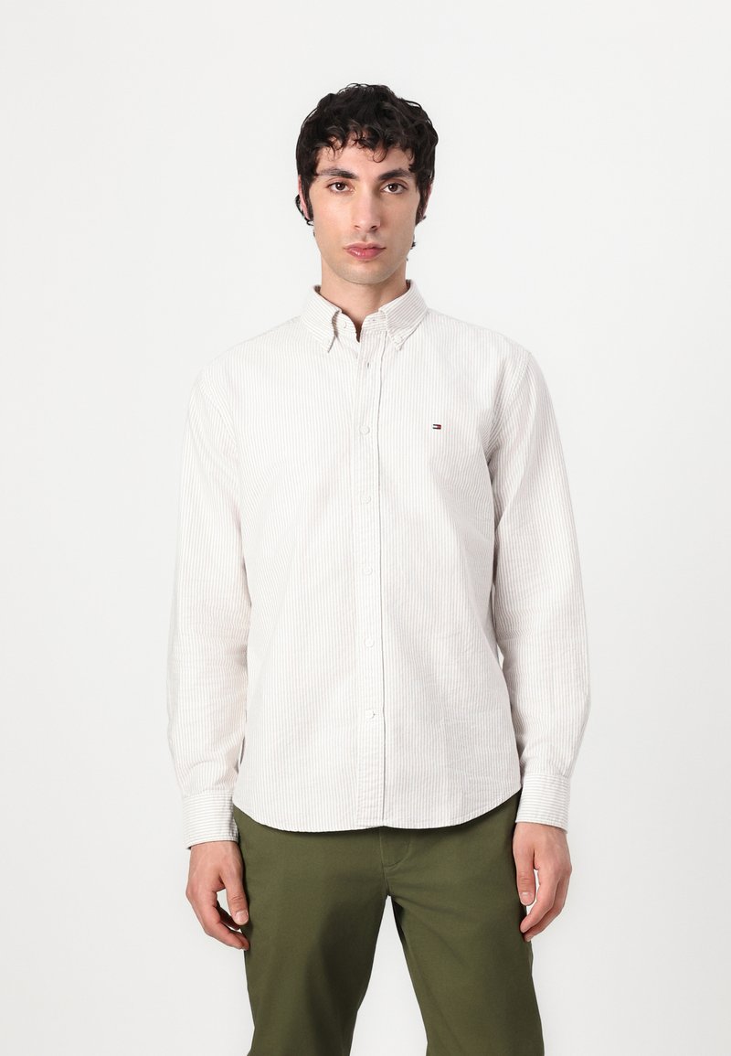 Camisa de botones blanca y gris claro a rayas, con un pequeño logo en el pecho, que presenta mangas largas y un collar estándar.