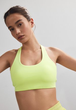 Junge Frau mit zurückgebundenem dunklem Haar, trägt einen hellgrünen Sport-BH und passende Shorts vor einem schlichten hellen Hintergrund.