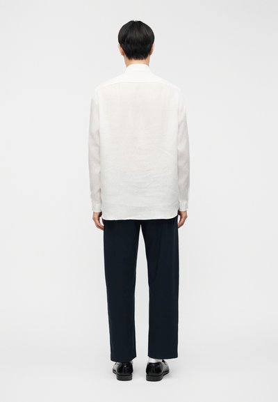 Chemise en lin blanc à coupe décontractée, manches longues, détail de la pièce d'épaule arrière, associée à un pantalon bleu marine foncé et des chaussures noires.