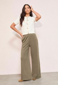 Balts īso piedurkņu adīts tops ar zelta pogām, savienots ar plašiem olīvzaļa ribota auduma biksēm. Gludas tekstūras un brīvs piegriezums.