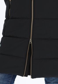 Schwarze gesteppte Jacke mit zweiseitigem Messing-Reißverschluss, unten teilweise geöffnet, getragen über blauen Jeans.