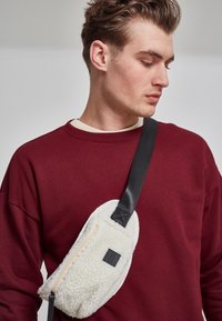 Fleece crossover bag i off-white, med glidelås og svart stropp. Designet er ovalt med en strukturert overflate og små logo-detaljer.