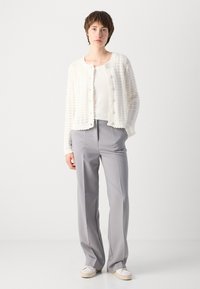 Weißer gestrickter Cardigan mit strukturiertem Muster, rundem Ausschnitt und Knopfverschluss, getragen über einem weißen Oberteil und hellgrauen weiten Hosen.