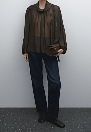 Person trägt eine schwarze, durchsichtige Bluse mit langen Ärmeln, dunkelblaue Jeans, schwarze Schuhe und hält eine braune, mit Nieten besetzte Handtasche vor einer schlichten grauen Wand.