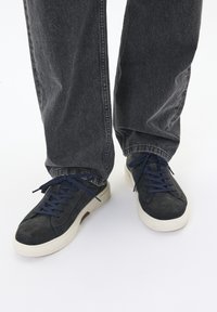 Tumedate suede sneakerid tekstuursed ülemise osaga, millel on valge kummist tall ja tumesinised paelad. Kandmine tumedate hallide lõdva lõikega teksadega.