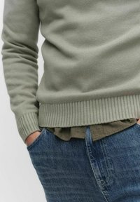 Ljusgrön stickad tröja med ribbade muddar och nedkant, lager på lager över en mörkgrön skjorta, tillsammans med blå denimjeans.