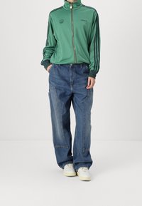 Grön idrottsjacka med dragkedja och svarta ränder, i kombination med lösa blå denimjeans med stora fickor och subtilt blekt mönster. Vita sneakers.