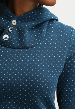 Nainen sinisessä pilkullisessa hupparissa, jossa on kolme koristeellista nappia lähellä kaulaa, näkyvissä ylävartalo ja osa kasvoista.