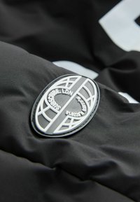 Le tissu noir présente une patch texturé avec un design de globe blanc et un lettrage en relief. Le matériau est lisse avec un léger éclat.