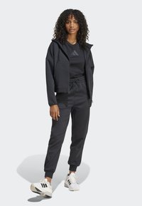 Svart treningstøy bestående av en hettegenser med glidelås og cropped design samt smale bukser, begge i et mykt materiale. Kombinert med hvite joggesko og svarte detaljer.