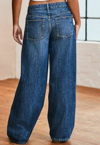 Leveled blue denim jeans with a faded texture, featuring two back pockets and a high waist, displayed on a wooden floor background.