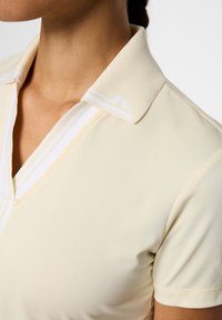 Jasnożółta koszulka polo z teksturowanym kołnierzykiem. Posiada białe paski i dekolt w serek. Gładka tkanina z krótkimi rękawami.